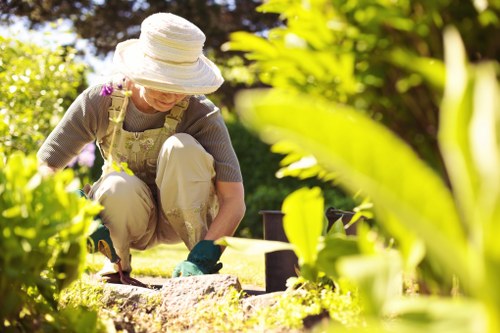 A woman in a sun hat and gardening gloves is kneeling on a well-maintained lawn in a front garden, tending to a flower bed border with dark soil. She is dressed in light-colored trousers and a beige apron, using a small hand tool, possibly a trowel, to work on the garden. Surrounding her are lush green plants and shrubbery, with a backdrop of trees and bushy foliage that filters sunlight, creating a bright outdoor environment. To her right, there are black garden pots placed on the grass, suggesting ongoing planting activity. The garden features a mix of cultivated flower beds and natural grass, with a defined edge separating the planting area from the lawn. The grass appears dense and healthy, exhibiting a rich, green hue typical of well-maintained outdoor spaces in Rotherhithe. The scene captures a peaceful gardening moment, reflecting services related to lawn care, planting, and general outdoor maintenance, emphasizing natural textures, vibrant colors, and a tidy, inviting landscape suitable for residential outdoor spaces managed by local landscapers.