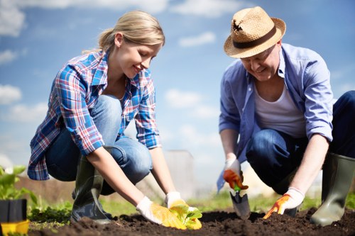 A young girl and an older woman are working together in a well-maintained garden, planting and tending to the soil. The girl is wearing a blue, red, and white checkered shirt, and both are dressed in garden-appropriate clothes with gloves. The garden features a lush, green lawn in the background, alongside neatly bordered flower beds with rich, dark soil that they are actively planting in. The scene is set outdoors on a bright, sunny day with a clear blue sky, likely in a residential garden in Rotherhithe or nearby South London, emphasizing sustainable gardening practices. The woman is wearing a straw hat for sun protection, and the garden environment appears organized and healthy, promoting eco-friendly outdoor maintenance aligned with local gardening services such as those provided by Landscapers Rotherhithe. Natural tones of green, brown, and the vibrant colors of the plants highlight the nurturing aspect of gardening, supporting sustainable and recycling initiatives in outdoor spaces.