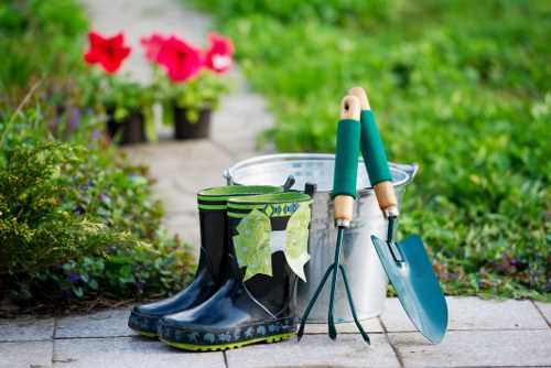 A pair of black and green gardening rubber boots placed on a paved pathway, with a pair of gardening tools including a hand trowel and pruning shears resting against a metal bucket. In the background, there are flower pots with vibrant pink and red petunias, set amidst a lush, well-maintained garden with green grass and various plants. The scene suggests an outdoor gardening activity, with natural daylight illuminating the area, and the garden appears organized and suitable for maintenance services offered by Landscapers Rotherhithe in the local area near Rotherhithe, London, highlighting practical gardening tools and bright floral accents within a landscaped garden environment.