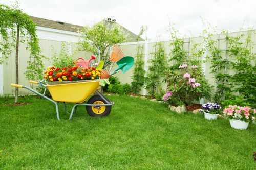 A well-maintained backyard garden featuring a lush, green lawn with evenly cut grass in the foreground. In the middle of the yard, a yellow wheelbarrow is filled with colorful garden flowers, including reds, oranges, and yellows, and is positioned beside a set of garden tools such as a watering can and a small shovel. Along the right side of the garden, there is a flower bed bordered by a low brick edge, with blooming plants in shades of pink, purple, and white, some in pots and others directly planted in the soil. The garden is enclosed by a white wooden fence, behind which tall green climbing plants and bushes create a natural privacy screen. In the background, a small tree with light green foliage provides additional shade and structure. The overall scene is bathed in natural daylight with an overcast sky, suggesting a calm and mild weather typical of outdoor garden maintenance in Rotherhithe, London. This setting reflects professional garden and landscaping services, focusing on planting, lawn care, and outdoor beautification within a suburban environment.