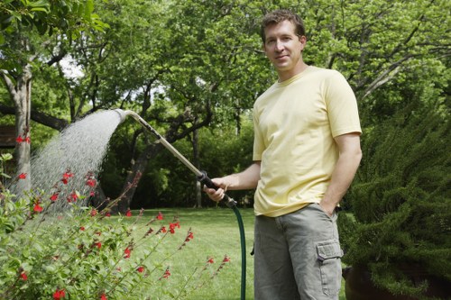 A man standing in a well-maintained garden, holding a garden hose that is spraying water over flowering plants and greenery. The garden features a lush, green lawn with a variety of shrubs and flower beds bordered by neatly trimmed bushes. In the background, there are tall trees providing shade, and the scene appears to be outdoors on a bright day with natural light. The overall setting highlights outdoor gardening and lawn care activities, reflecting professional landscaping and garden maintenance services offered by landscapers in Rotherhithe, with natural tones of green, red, and earth colours present in the image.