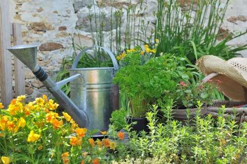 A vibrant and well-maintained garden scene featuring a variety of plants and gardening elements. In the foreground, there are bright yellow and orange flowering plants, adding vivid colour to the space. Behind these, lush green foliage and shrubs grow densely, with some taller plants and grasses providing vertical interest. A weathered metal watering can is placed among the plants, indicating active garden care. To the right, a straw garden hat rests on the edge of a flower bed or decorative container, suggesting recent gardening activity. The garden is bordered by a low stone wall, creating a rustic backdrop, and a wooden garden feature or trellis is partially visible on the left side. The overall scene is outdoors, likely in a small-to-medium-sized landscaped yard in Rotherhithe, with natural light illuminating the space, highlighting the variety of textures and tones from the natural greens and earth tones to the cheerful blooms. This image exemplifies typical gardening and outdoor maintenance practices that a professional company like Landscapers Rotherhithe might support, emphasizing the importance of plant care, watering, and garden organisation typical of local residential gardens in the South East London area, specifically around postcode SE16.