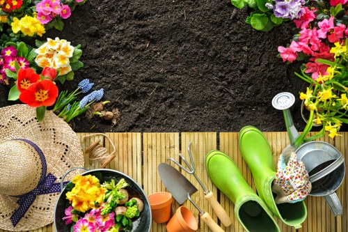 An overhead view of a neatly arranged garden corner at a property in Rotherhithe, featuring a wooden decking area bordered by a variety of colorful flowering plants including primroses, pansies, and hyacinths along the edge of freshly turned dark soil. The garden incorporates a lush green lawn adjacent to the flower borders, with garden tools such as a trowel, watering can, and gardening gloves neatly placed on the decking. A large straw sun hat with a purple ribbon rests among the plants, adding a touch of outdoor leisure. The scene demonstrates well-maintained landscaping suitable for outdoor gardening services offered by Landscapers Rotherhithe, capturing the natural tones of the soil, vibrant blossoms, and the textured surface of the wooden decking under natural daylight, ideal for enhancing local garden care and landscaping projects in the area.