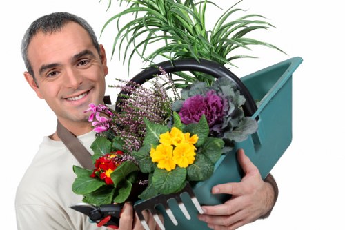 The image shows a man smiling and holding a green plastic garden watering can filled with a variety of potted plants and flowers in vibrant colours, including yellow, purple, red, and pink. Behind him, there is a tall, lush green plant with multiple slender leaves. The scene is set against a plain white background, emphasizing the gardening and outdoor maintenance theme associated with Rotherhithe local landscaping services. The arrangement of the plants suggests a focus on garden planting and care, aligning with professional gardening, lawn maintenance, and planting services offered by companies like Landscapers Rotherhithe. The overall composition highlights the joy and vibrancy of outdoor garden care, suitable for a landscaping service webpage promoting garden planting and maintenance expertise in the Rotherhithe area, near London, with natural lighting enhancing the fresh, healthy appearance of the plants.