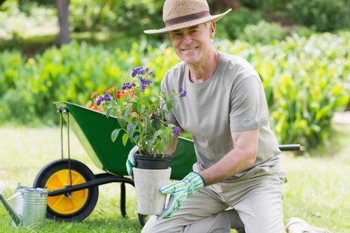 A gardener, dressed in a light grey t-shirt, beige trousers, a straw hat, and gardening gloves, is kneeling on well-maintained green grass in a backyard or garden area. He is holding a potted plant with purple flowers, which is freshly dug from the soil. Behind him, there is a bright yellow wheelbarrow with a black handle and wheel, filled with gardening supplies, set on a lush lawn bordered by a hedge or shrubbery. In the background, tall leafy trees and dense greenery provide natural shade and a vibrant outdoor environment typical of Rotherhithe or similar South London gardens. The scene captures a clear, sunny day, showcasing natural tones of green, purple, and earthy browns, emphasizing outdoor maintenance and gardening activities. This image exemplifies professional gardening and landscaping services offered by Landscapers Rotherhithe, highlighting plant care and garden enhancement in a typical residential garden setting, connecting visually to local outdoor spaces and garden improvement projects.