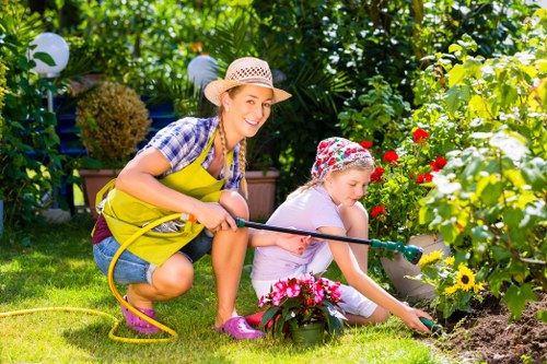In a well-maintained backyard garden, a woman and a young girl are engaged in outdoor gardening activities. The woman, wearing a straw hat, a blue and white checkered shirt, a bright yellow gardening apron, and pink waterproof shoes, is crouched down on the lush green lawn holding a yellow garden hose, smiling at the camera. The girl, dressed in light clothing and a floral bucket hat, is focused on planting or tending to flowering plants in a flower bed bordered by a stone walkway. The flower bed features vibrant red, yellow, and pink blooms, adding bright natural colours to the scene. Behind them, dense foliage, including a hedge, leafy trees, and potted plants, creates a lush, private outdoor environment. The garden is illuminated by natural sunlight, creating a cheerful and inviting atmosphere, indicative of a typical landscaped suburban space in the Rotherhithe area. The scene reflects outdoor maintenance and gardening care provided by local experts like Landscapers Rotherhithe, emphasizing a well-tended garden setting with a focus on flower cultivation, lawn care, and outdoor enjoying activities.