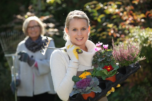 A young woman with blonde hair, wearing a white sweater and yellow gardening gloves, is standing outdoors in a garden during daylight, holding a black tray filled with various colourful flowering plants and greenery, with a smiling older woman in the background also dressed for gardening, wearing glasses and a scarf, in front of a lush garden with trees and shrubbery. The scene suggests a garden maintenance or planting activity, with natural sunlight illuminating the vibrant tones of the flowers and foliage, highlighting the well-kept lawn and garden features, and depicting a typical outdoor gardening environment in Rotherhithe, aligned with local gardening services by Landscapers Rotherhithe.
